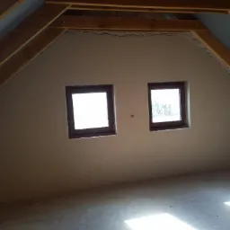 Poddasze z dwoma oknami po nałożeniu jasnego tynku maszynowego na ściany, widoczne drewniane elementy konstrukcji dachu.