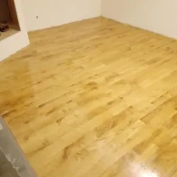 Świeżo ułożona, lakierowana podłoga z paneli w pustym pomieszczeniu z białymi ścianami, widoczne niedoskonałości w wykończeniu przy ścianie.