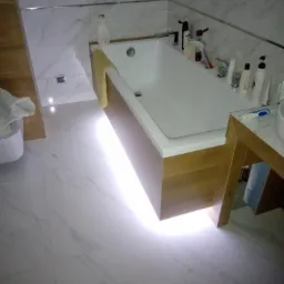 Nowoczesna łazienka z podświetlaną wanną obudowaną drewnem, białe marmurowe płytki na ścianach i podłodze, minimalistyczna armatura, widok na umywalkę z drewnianym blatem.