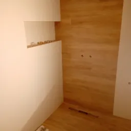 Wykończona łazienka z brązowymi płytkami imitującymi drewno na ścianie prysznica, białe płytki na pozostałych ścianach, widoczny odpływ liniowy i armatura na półce.