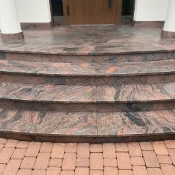 Granitowe schody wejściowe z zaokrąglonymi stopniami, z kolumnami po bokach i kostką brukową u dołu, z widocznym wzorem na kamieniu.