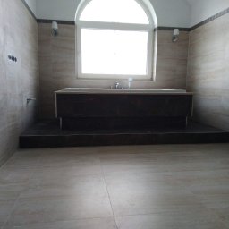 J&L Comfort - Beżowa łazienka z wanną na podwyższeniu, wykończona płytkami imitującymi trawertyn, z oknem w kształcie łuku i minimalistycznymi kinkietami.