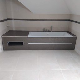 J&L Comfort - Nowoczesna zabudowa wanny z ciemnoszarą mozaiką i jasnymi płytkami pod skosem dachu, widoczny fragment osoby w drzwiach.