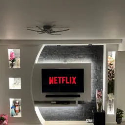 Elegancka ściana telewizyjna z podświetlanymi wnękami, wykończona szarą okładziną imitującą kamień, z dekoracjami w postaci kwiatów i alkoholu, oświetlona nowoczesną lampą sufitową.