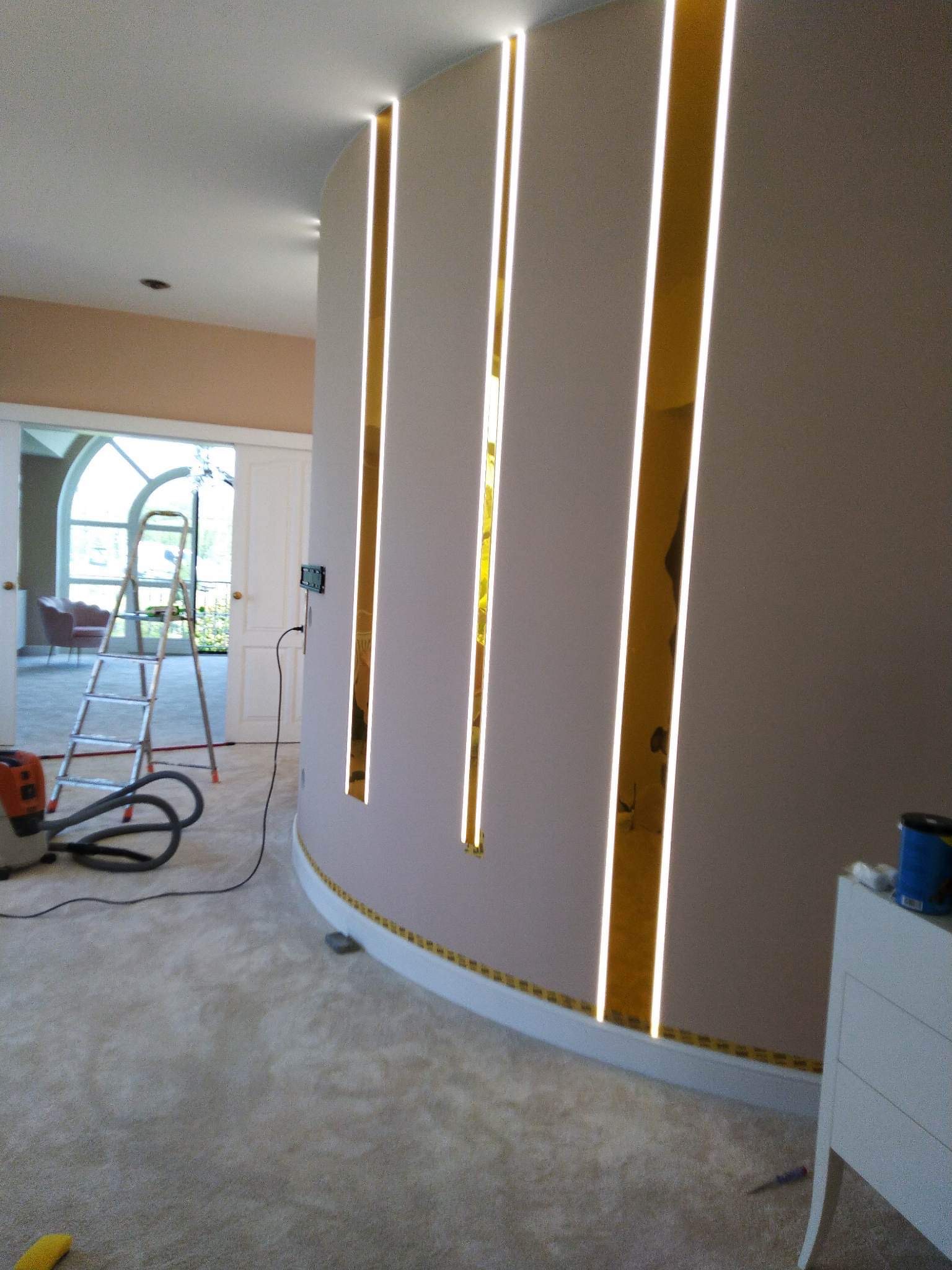 Zaokrąglona ściana z trzema pionowymi wnękami podświetlonymi ledowym światłem, widoczna drabina, odkurzacz i meble w tle, zabezpieczona taśmą malarską listwa przypodłogowa.