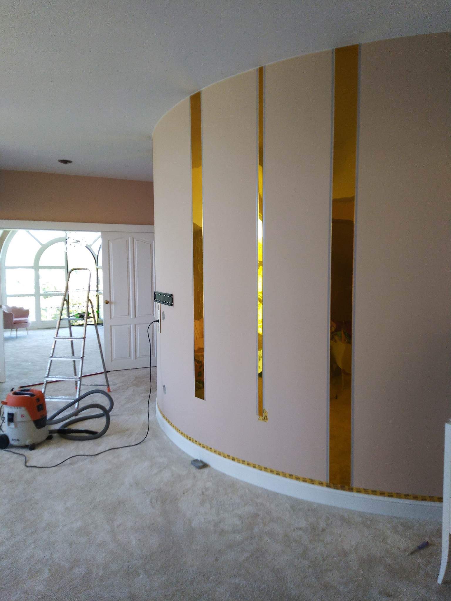 Zaokrąglona ściana z beżową farbą i złotymi, pionowymi wstawkami, zabezpieczona taśmą malarską przy listwie podłogowej, w tle drabina malarska i odkurzacz