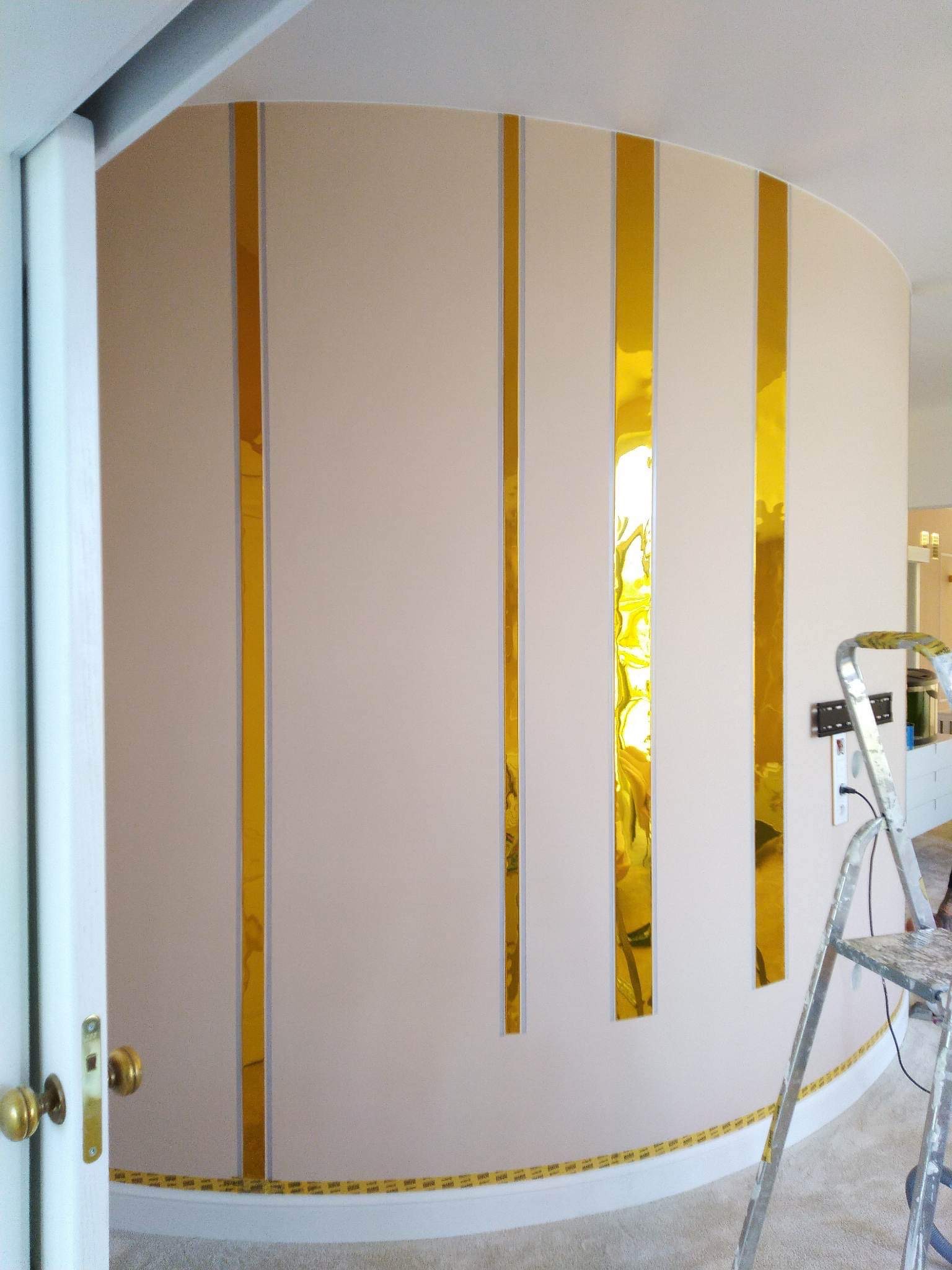Zakrzywiona ściana z pionowymi, złotymi, lustrzanymi paskami, zabezpieczona taśmą malarską przy podłodze, z widoczną drabinką malarską i otwartymi drzwiami w tle.