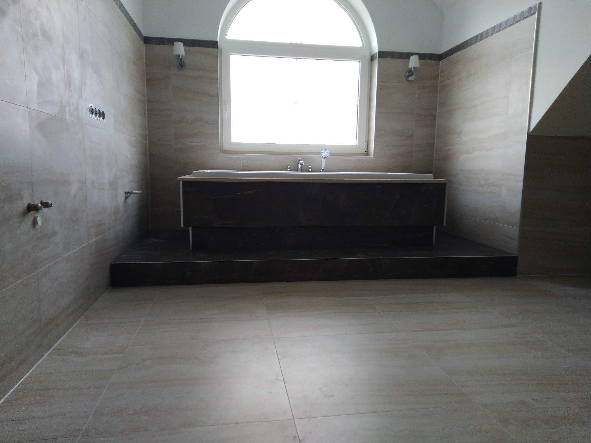 Beżowa łazienka z wanną na podwyższeniu, wykończona płytkami imitującymi trawertyn, z oknem w kształcie łuku i minimalistycznymi kinkietami.