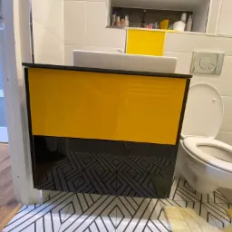 Ż&oacute;łto-czarna szafka pod umywalkę z błyszczącymi frontami, wisząca nad podłogą w geometryczne wzory, obok toaleta i wnęka z kosmetykami.