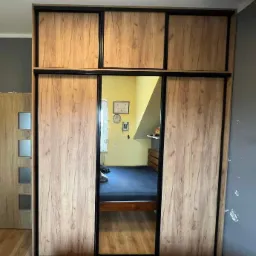 Wrocław: Szafa wnękowa z przesuwnymi drzwiami z okleiną imitującą jasne drewno i czarnymi obramowaniami, z lustrem w centralnej części, odzwierciedlającym wnętrze pokoju z łóżkiem i żółtą ścianą.