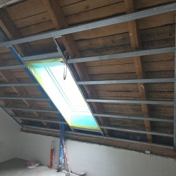 Remo-Dom Usługi Remontowo-Budowlane Krzysztof Żądełek - Skośne poddasze w trakcie adaptacji: drewniana konstrukcja dachu z metalowym stelażem, okno dachowe zabezpieczone folią, ściany częściowo otynkowane, widoczne instalacje.