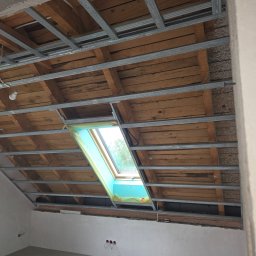 Remo-Dom Usługi Remontowo-Budowlane Krzysztof Żądełek - Montaż metalowej konstrukcji pod płyty G-K na drewnianym poddaszu z oknem dachowym. Widoczne profile i fragment ściany z instalacją elektryczną.