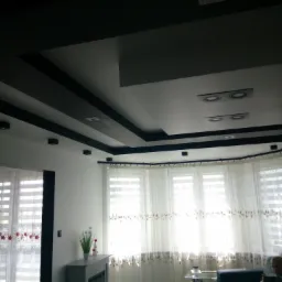 Nowoczesny salon z podwieszanym sufitem z czarnymi belkami, białymi ścianami, firanami w oknach i dekoracyjnym kominkiem elektrycznym.