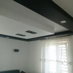 Nowoczesny salon z białym sufitem podwieszanym, czarnymi elementami dekoracyjnymi i oświetleniem punktowym, widok na okno z firanką i fragment sofy.