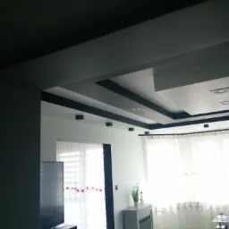 Nowoczesny salon z podwieszanym sufitem w odcieniach szarości, białymi ścianami i jasnymi zasłonami w oknach.