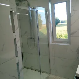 Nowoczesna kabina prysznicowa z deszczownicą, wykończona płytkami imitującymi marmur, z widokiem na pole kukurydzy za oknem.