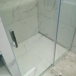 Kabina prysznicowa z transparentnego szkła i chromowanymi okuciami, wykończona białymi płytkami imitującymi marmur na ścianach i podłodze, z widocznym odpływem liniowym.