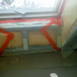 Montaż izolacji termicznej poddasza wokół okna dachowego, widoczne profile metalowe, folia paroizolacyjna z taśmą uszczelniającą Roto, fragment drabiny w dolnym rogu.