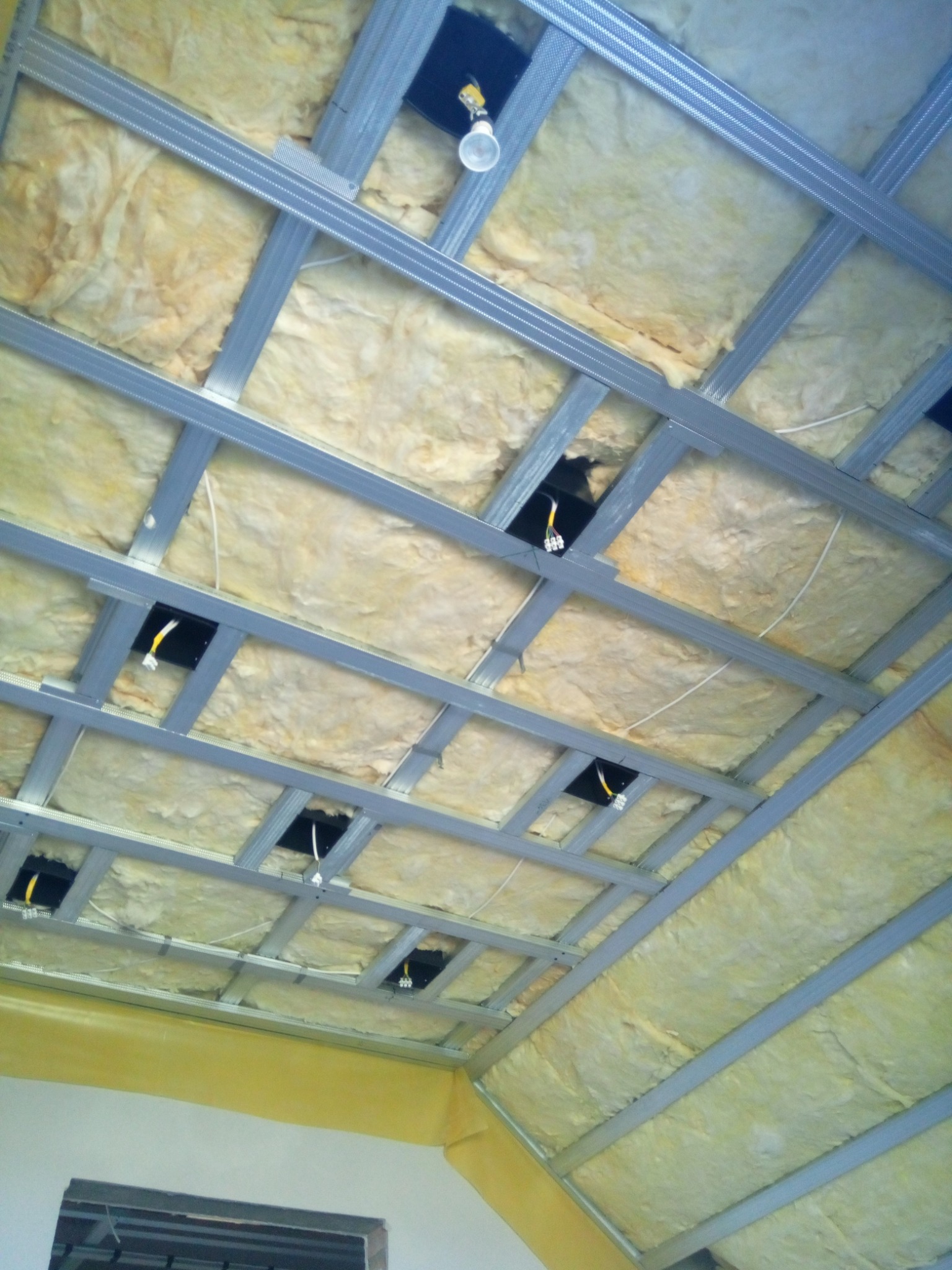 Skośny sufit z widoczną izolacją z wełny mineralnej, metalową konstrukcją nośną i otworami na oświetlenie z wystającymi przewodami elektrycznymi.