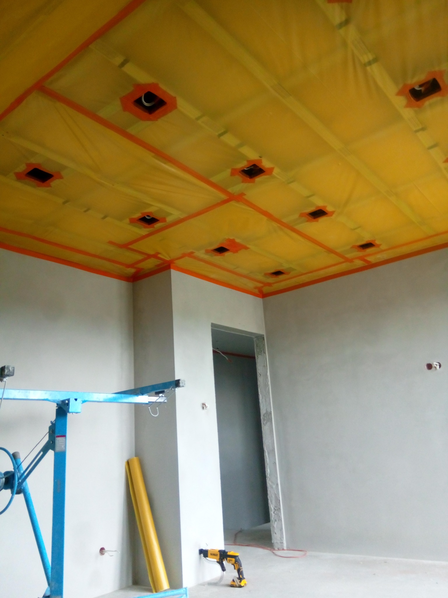 Sufit z żółtą folią paroizolacyjną, zabezpieczoną pomarańczową taśmą, przygotowany do montażu oświetlenia punktowego, widoczny podnośnik do płyt gipsowo-kartonowych i zwinięta folia