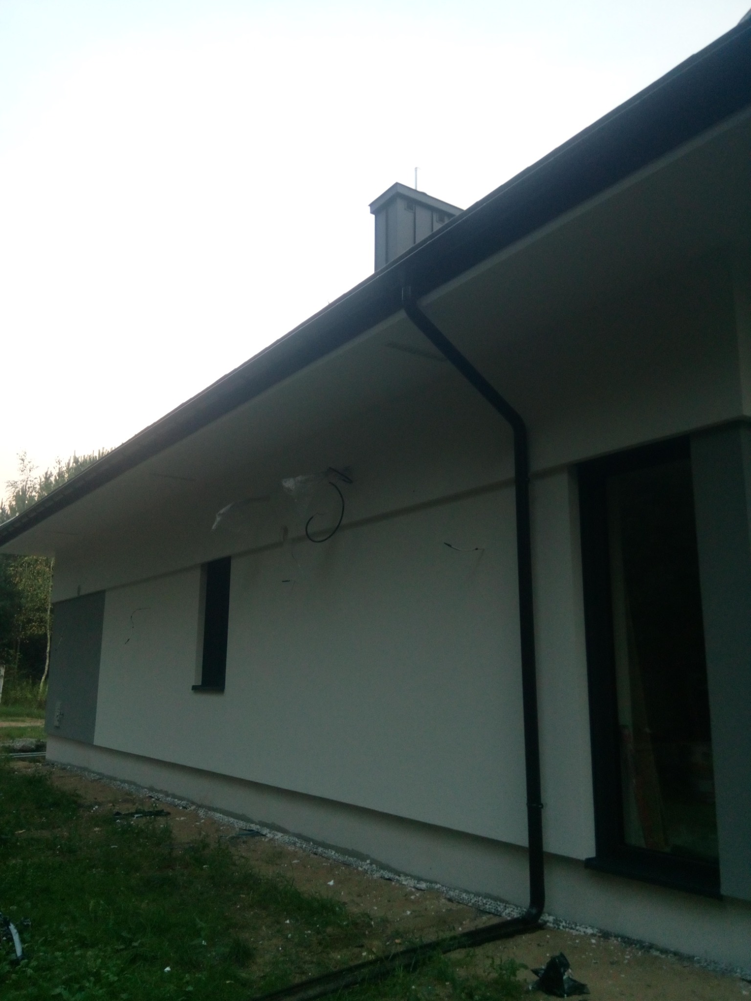 Ściana zewnętrzna domu jednorodzinnego w trakcie wykończenia, widoczne niedoskonałości na elewacji, czarna rynna spustowa i okno z ciemną ramą.