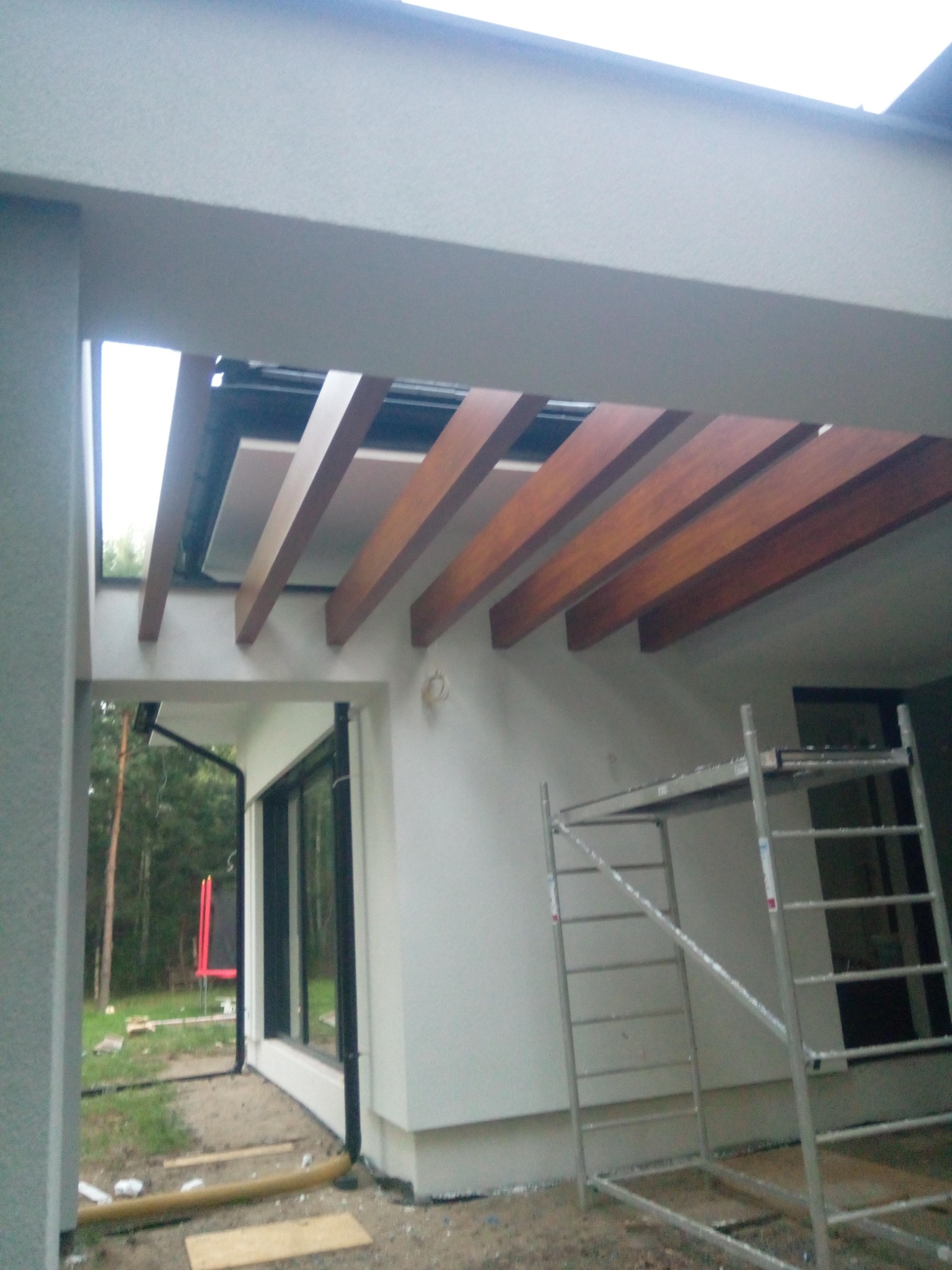 Nowoczesna pergola z drewnianymi belkami, widok od wewnątrz, z rusztowaniem w tle, częściowo ocieplony fragment dachu.