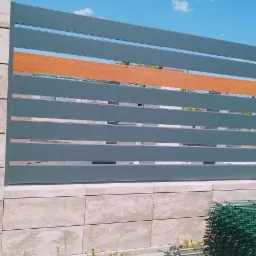 Szare, poziome przęsła z elementem dekoracyjnym imitującym drewno, osadzone między słupkami z jasnych, marmurkowych bloczk&oacute;w betonowych. U dołu widoczny stos zielonych paneli ogrodzeniowych.