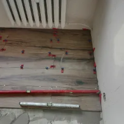 Układanie drewnopodobnych płytek na ścianie, widoczne poziomice i system poziomowania płytek, grzejnik na górze ściany.