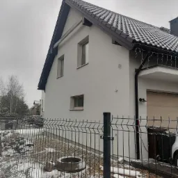 Świeżo otynkowany dom jednorodzinny z brązowym dachem w zimowej scenerii, widoczny fragment ogrodzenia panelowego i opady śniegu.