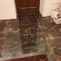 LinRom - Układanie płytek imitujących marmur na podłodze, widoczne pomarańczowe krzyżyki dystansowe i mata wejściowa przy drewnianych drzwiach.