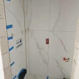 Wnętrze łazienki w trakcie wykańczania, widoczne białe płytki imitujące marmur, niebieska taśma malarska, puszka elektryczna i rury do armatury.