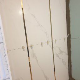 LinRom - Montaż białych płytek imitujących marmur z wąskimi złotymi listwami, użycie poziomowania krzyżykami na żyłce, widoczny stan surowy ściany i podłogi.