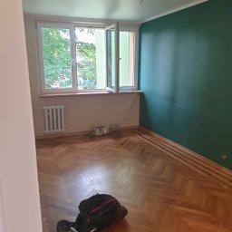 LinRom - Pokój w trakcie remontu: świeżo pomalowana ściana w odcieniach zieleni, parkiet w jodełkę, listwy przypodłogowe ułożone wzdłuż ściany, otwarte okno z widokiem na zieleń, grzejnik i torba...