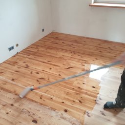 LinRom - Lakierowanie drewnianej podłogi z desek za pomocą wałka malarskiego; widoczne fragmenty pomalowanej i niepomalowanej powierzchni, skarpeta na stopie wykonawcy.
