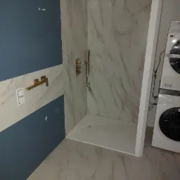 Nowoczesna kabina prysznicowa z marmurowymi płytami, złotą armaturą i niebieską ścianą w łazience z pralką.