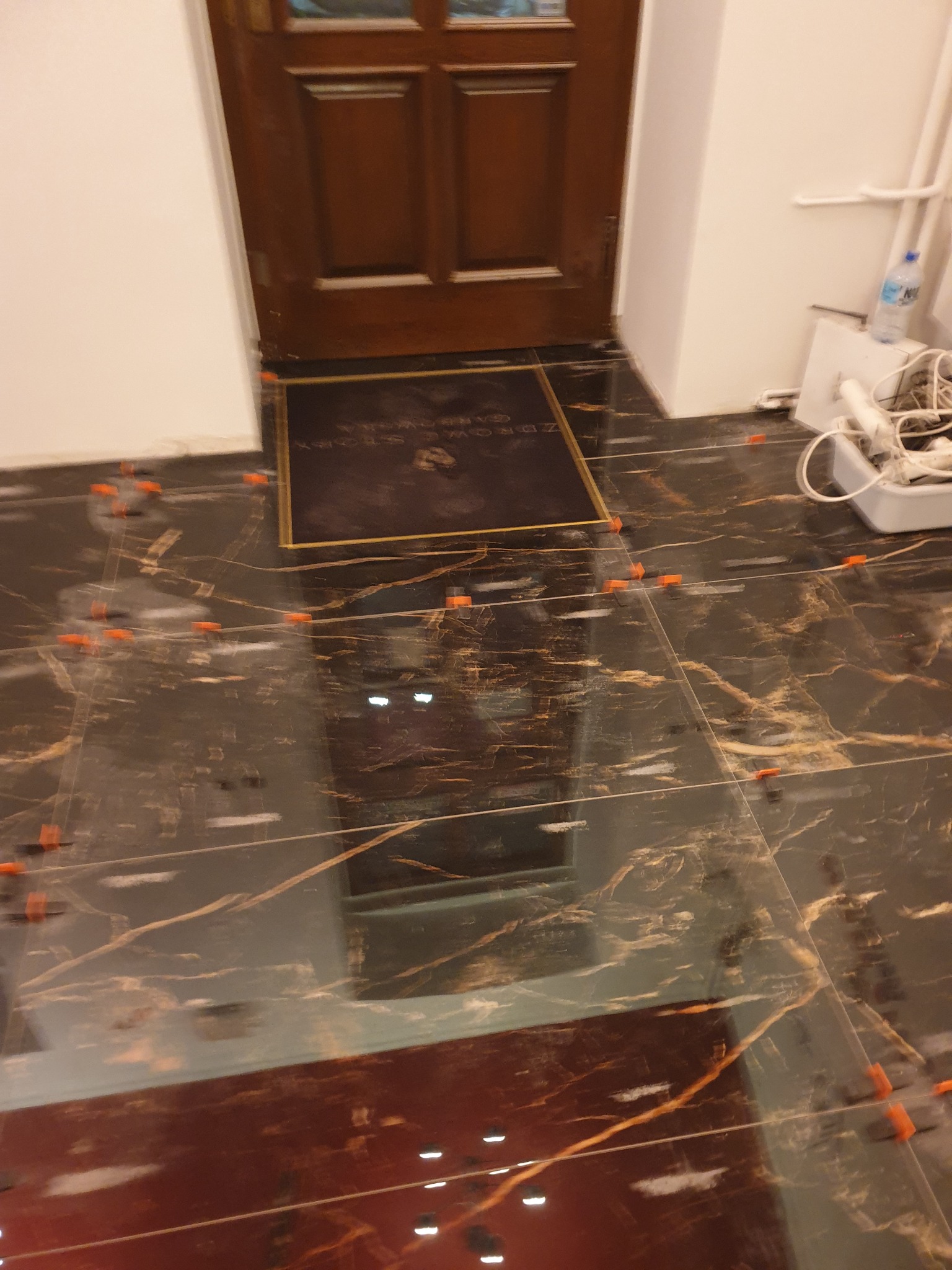 Układanie płytek imitujących marmur na podłodze, widoczne pomarańczowe krzyżyki dystansowe i mata wejściowa przy drewnianych drzwiach.