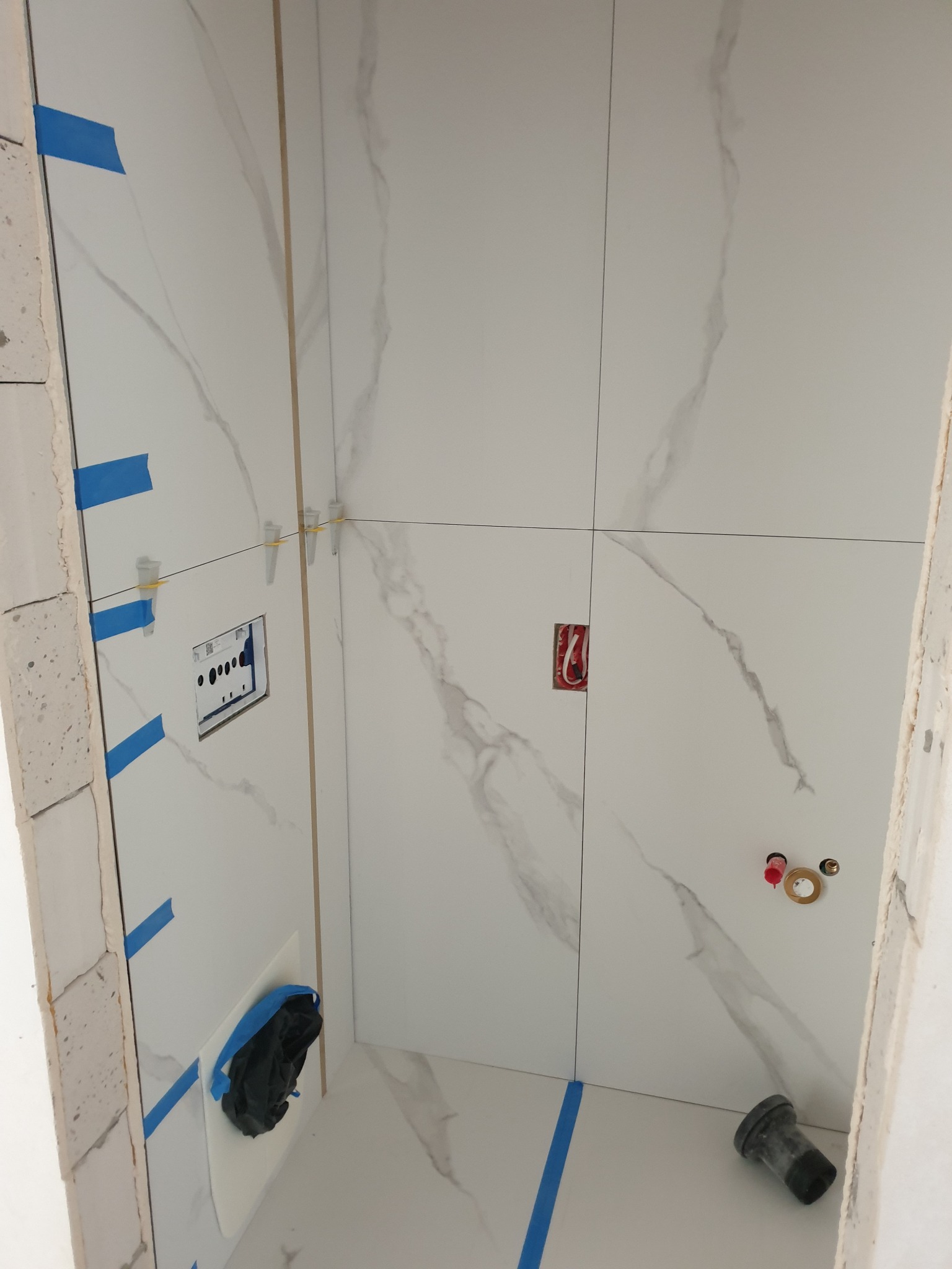Wnętrze łazienki w trakcie wykańczania, widoczne białe płytki imitujące marmur, niebieska taśma malarska, puszka elektryczna i rury do armatury.