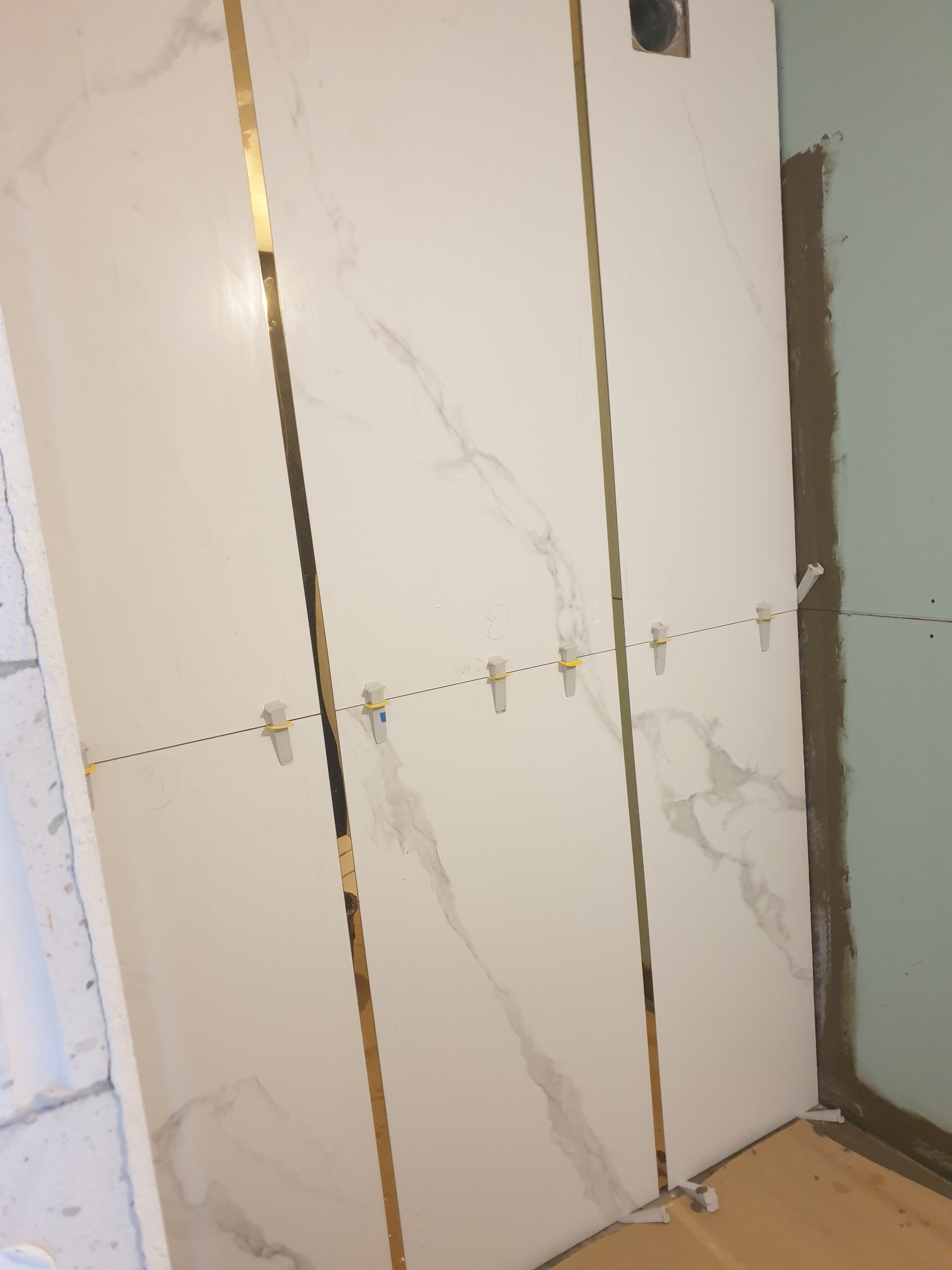 Montaż białych płytek imitujących marmur z wąskimi złotymi listwami, użycie poziomowania krzyżykami na żyłce, widoczny stan surowy ściany i podłogi.