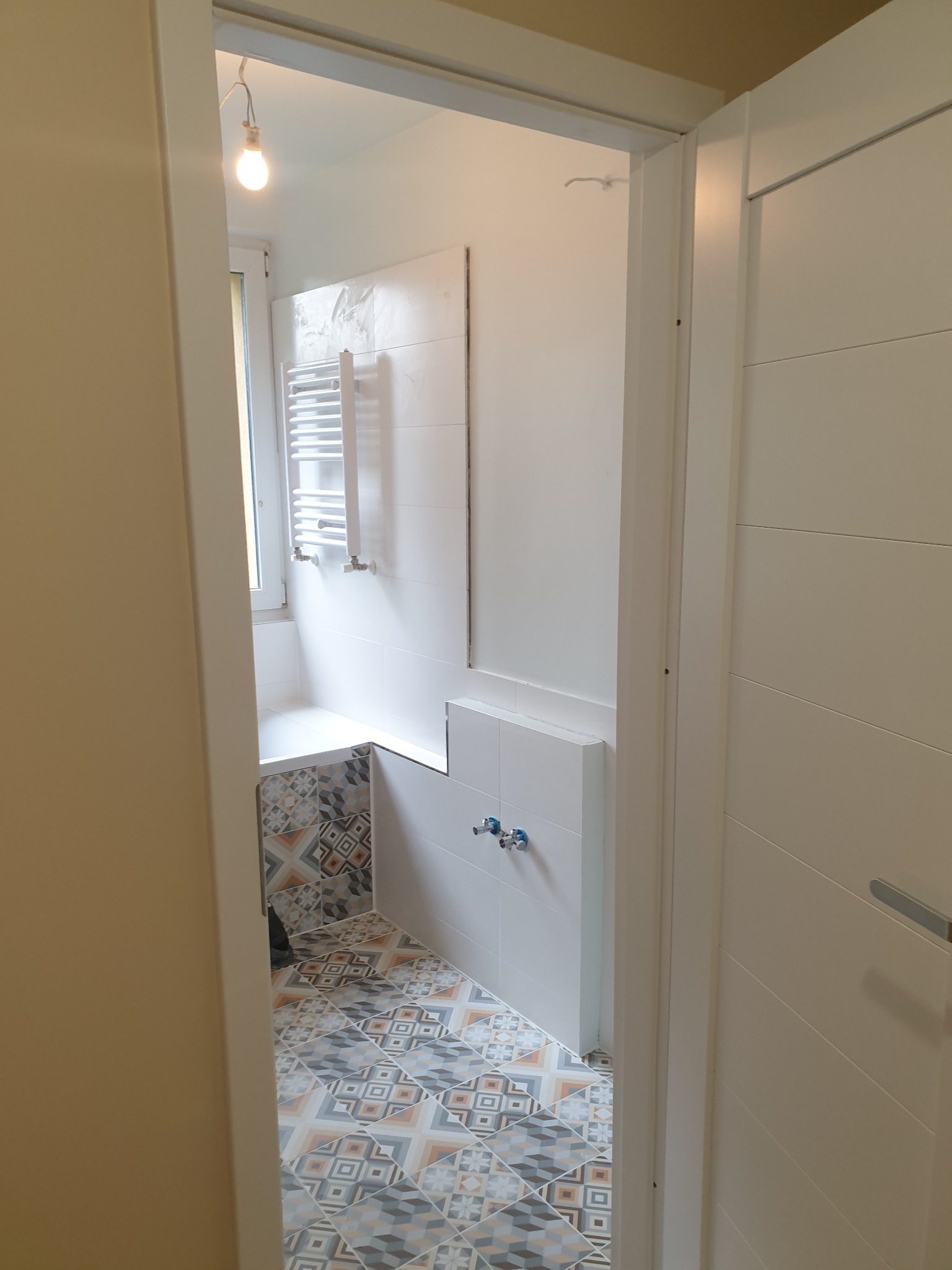 Widok z korytarza na wyremontowaną łazienkę z geometryczną mozaiką na podłodze i obudowie wanny, białe płytki na ścianach oraz grzejnik drabinkowy.