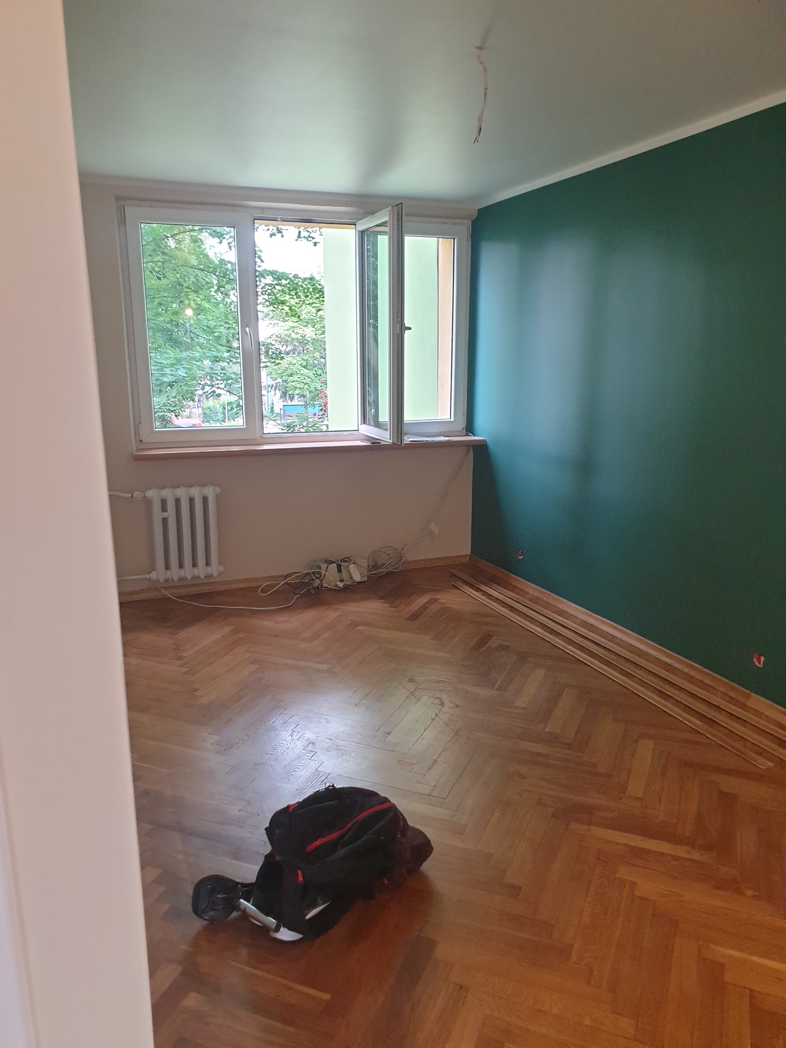 Pokój w trakcie remontu: świeżo pomalowana ściana w odcieniach zieleni, parkiet w jodełkę, listwy przypodłogowe ułożone wzdłuż ściany, otwarte okno z widokiem na zieleń, grzejnik i torba...
