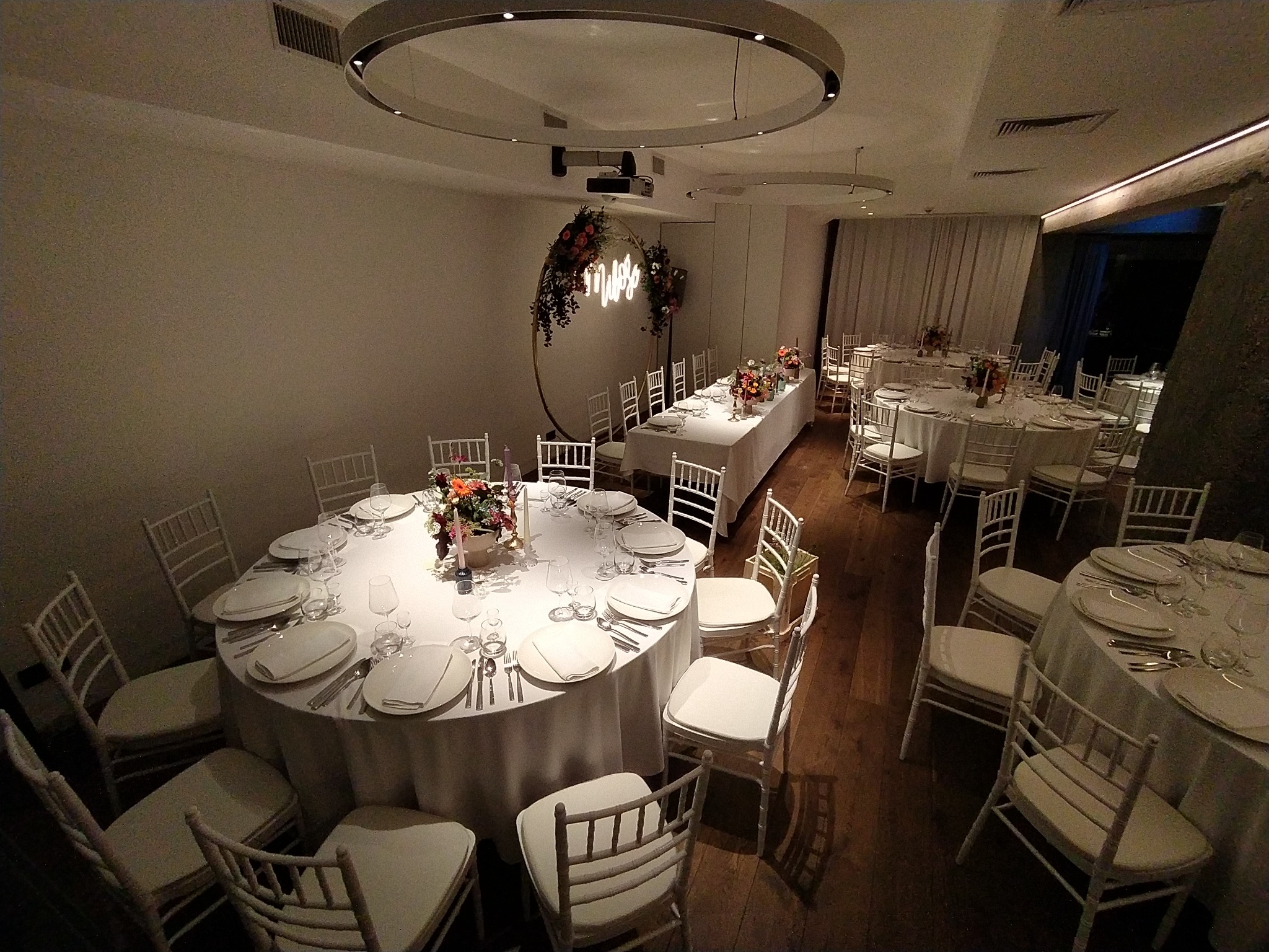 Sala bankietowa z okrągłymi stołami nakrytymi białymi obrusami, białymi krzesłami, dekoracjami kwiatowymi i podwieszanym oświetleniem w kształcie pierścienia. W tle dekoracja kwiatowa w kształcie...