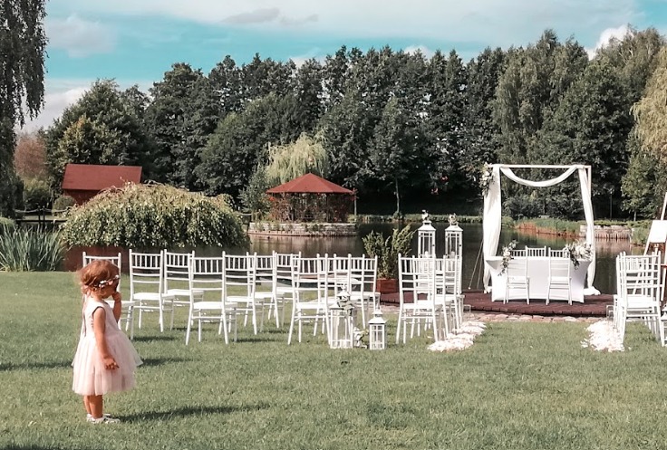 Uroczyste ustawienie krzeseł i dekoracji na trawie przed stawem z altaną, przygotowane na ceremonię ślubną na świeżym powietrzu, z małą dziewczynką w różowej sukience obserwującą przygotowania.