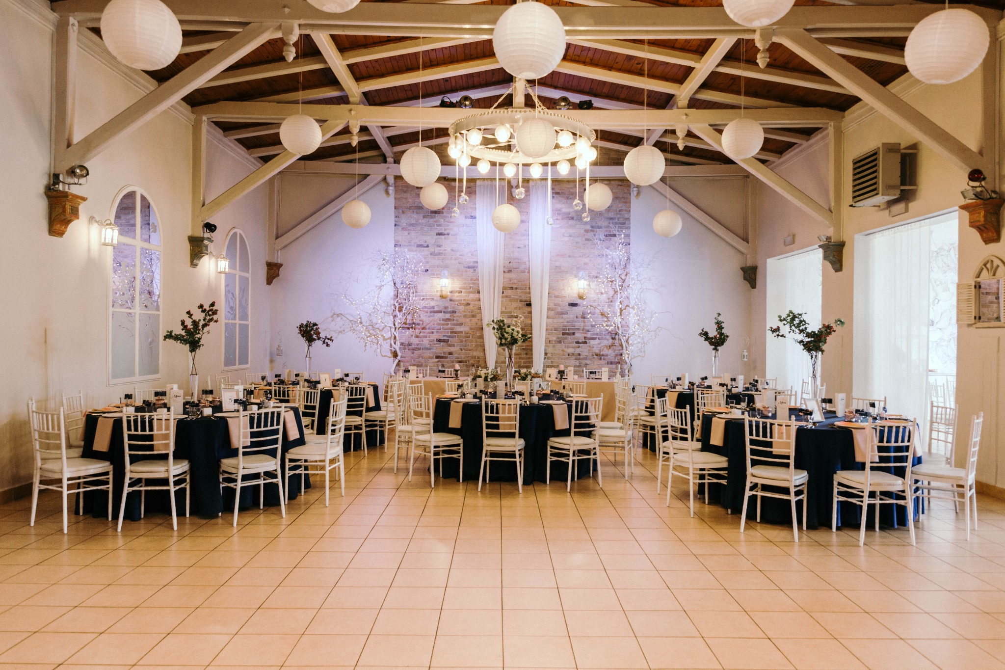 Sala bankietowa z okrągłymi stołami nakrytymi granatowymi obrusami i białymi krzesłami, dekoracje z papierowych lampionów i podświetlanych gałązek, ceglana ściana w tle.