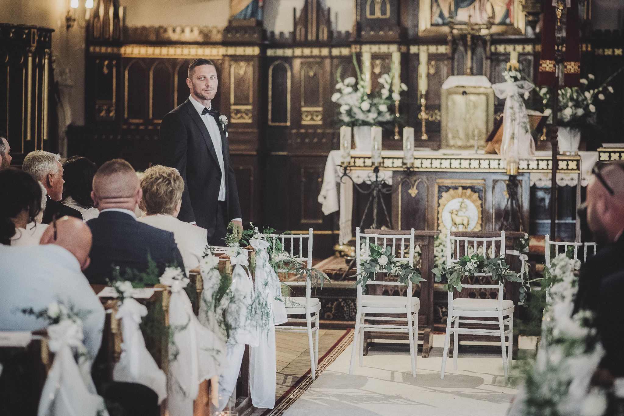 Mężczyzna w garniturze stoi w kościele, za nim rzędy białych krzeseł udekorowanych kwiatami i zielenią, w tle ołtarz i wierni.