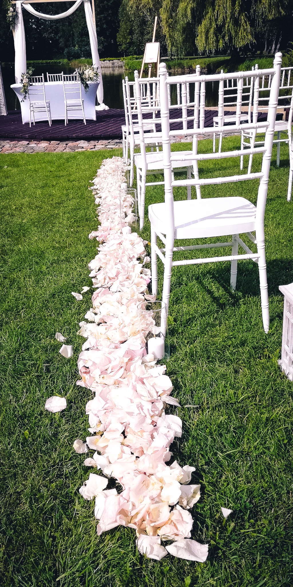 Uroczysta ścieżka z białych płatków róż prowadząca do białego, drewnianego stołu i krzeseł na tle zielonego trawnika i stawu, przygotowana na ceremonię ślubną.