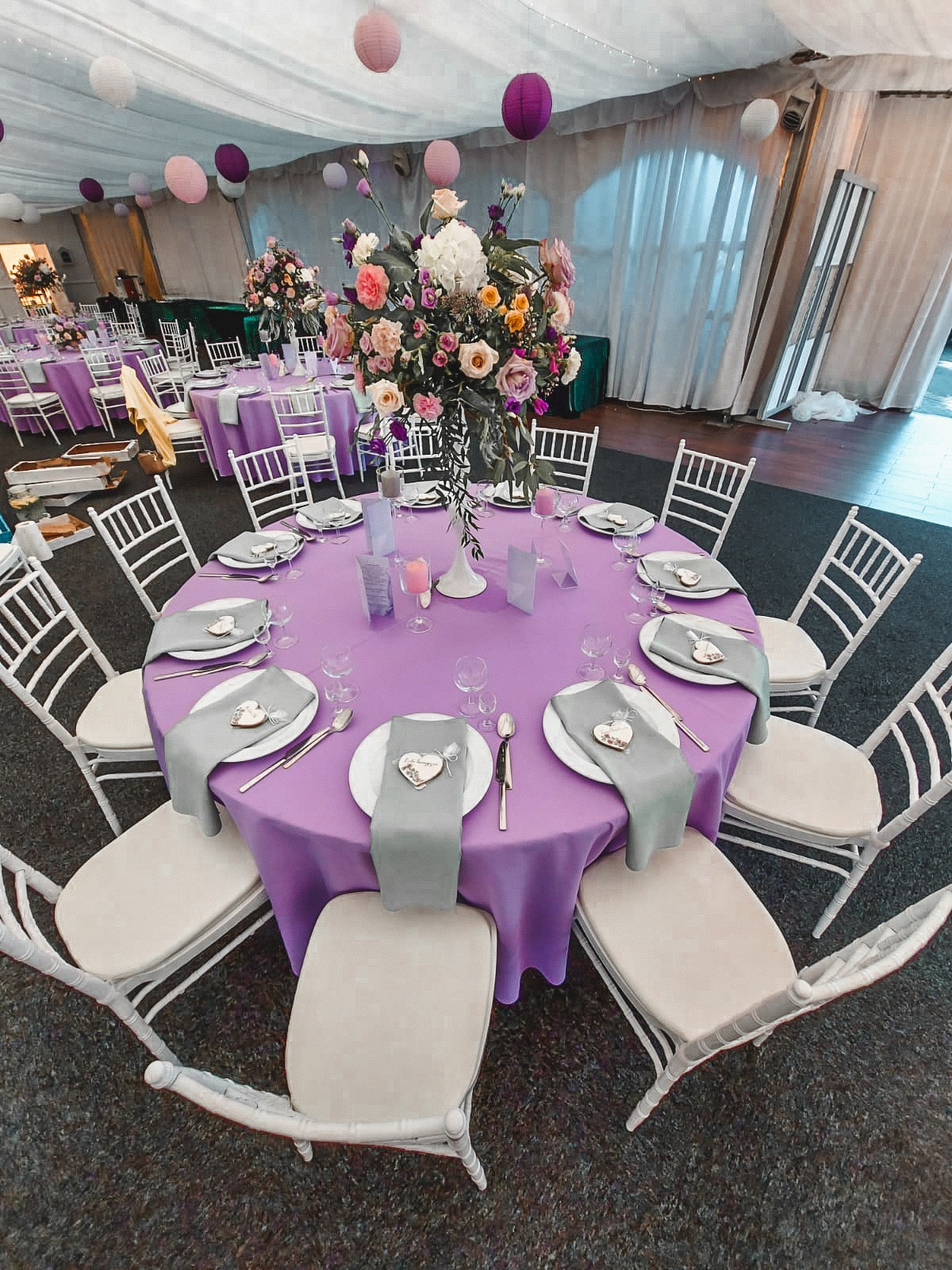 Uroczysty stół z fioletowym obrusem, białymi krzesłami i kwiatową dekoracją w Toruniu, przygotowany na przyjęcie w namiocie z lampionami.
