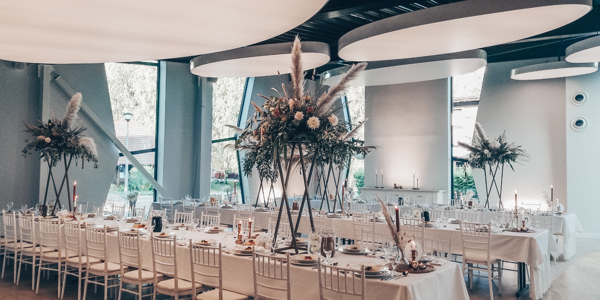 Uroczysta sala bankietowa z długimi stołami przykrytymi białymi obrusami, białymi krzesłami, wysokimi kompozycjami kwiatowymi z pampasami i świecami, oświetlona nowoczesnymi lampami sufitowymi.