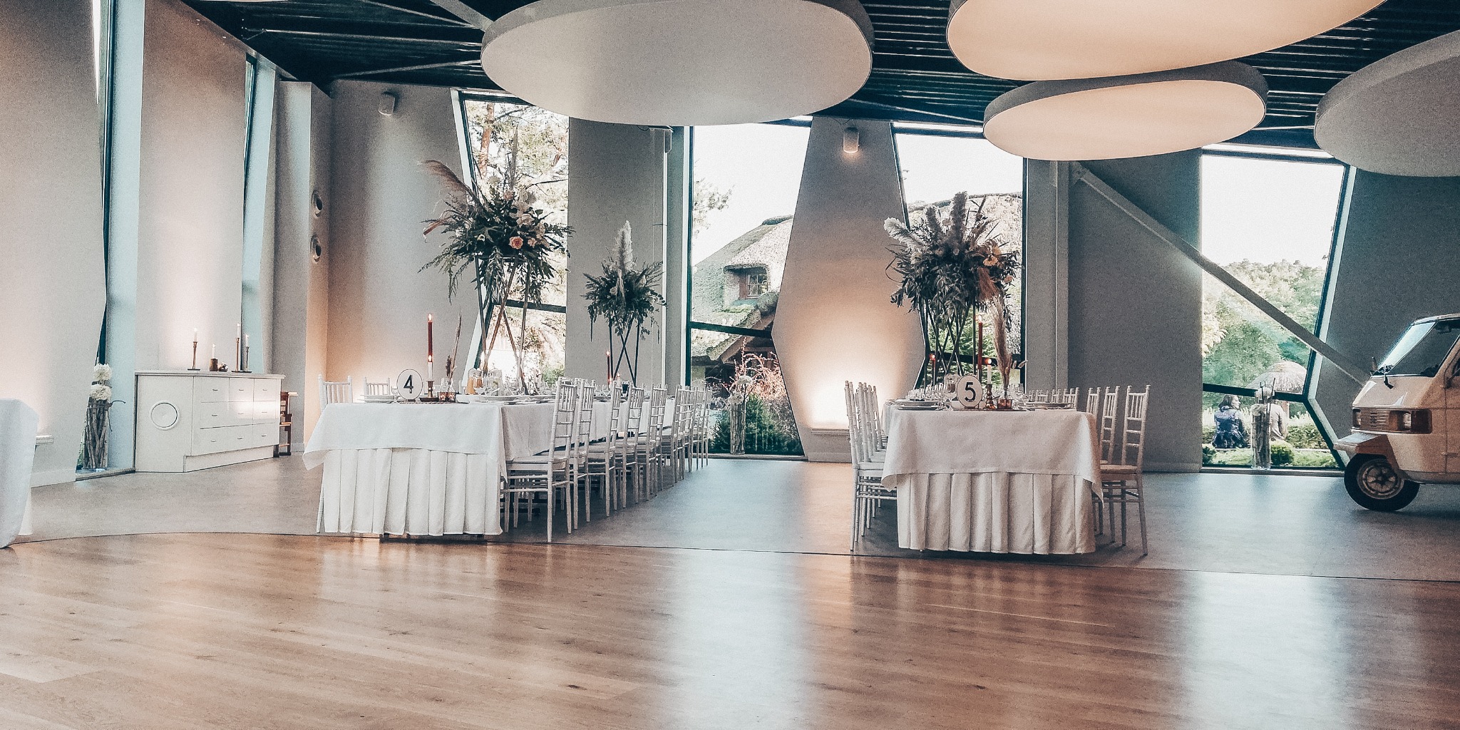 Uroczysta sala z dwoma nakrytymi stołami, wysokimi kompozycjami kwiatowymi i zapalonymi świecami, widok na zewnątrz przez okna o nietypowym kształcie, na zewnątrz stoi trójkołowy pojazd Ape.