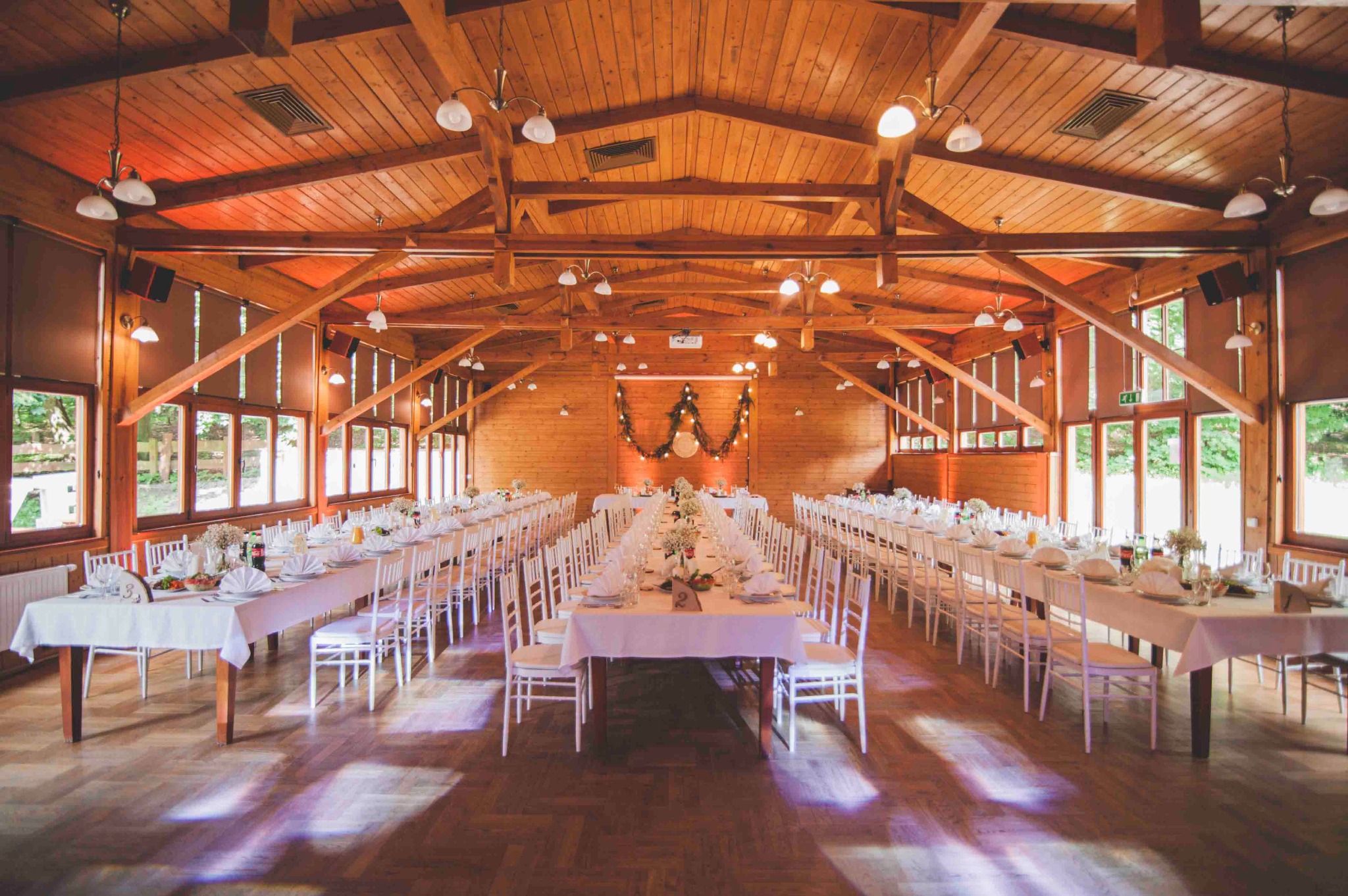 Sala weselna z drewnianą konstrukcją dachu, długie stoły z białymi obrusami i krzesłami, dekoracje kwiatowe i girlanda świetlna na ścianie, parkiet z refleksami światła.