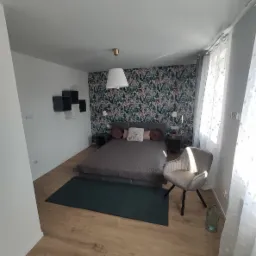 Sypialnia z łóżkiem kontynentalnym, tapetą w motywy roślinne, drewnianą podłogą i krzesłem w stylu retro, oświetlona wiszącą lampą z abażurem.